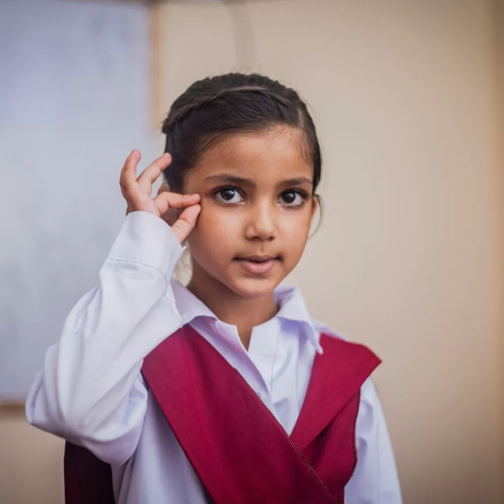 Girl in a white shirt with a red sash poses, making an OK gesture near her eye and looking at the camera.