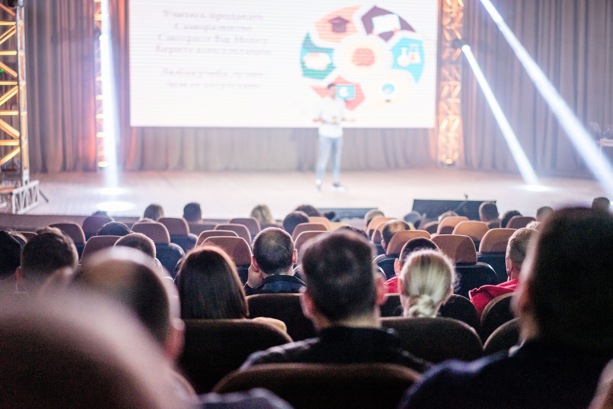 Speaker on stage giving a presentation to a seated audience in a theater with a large screen behind him and stage lighting