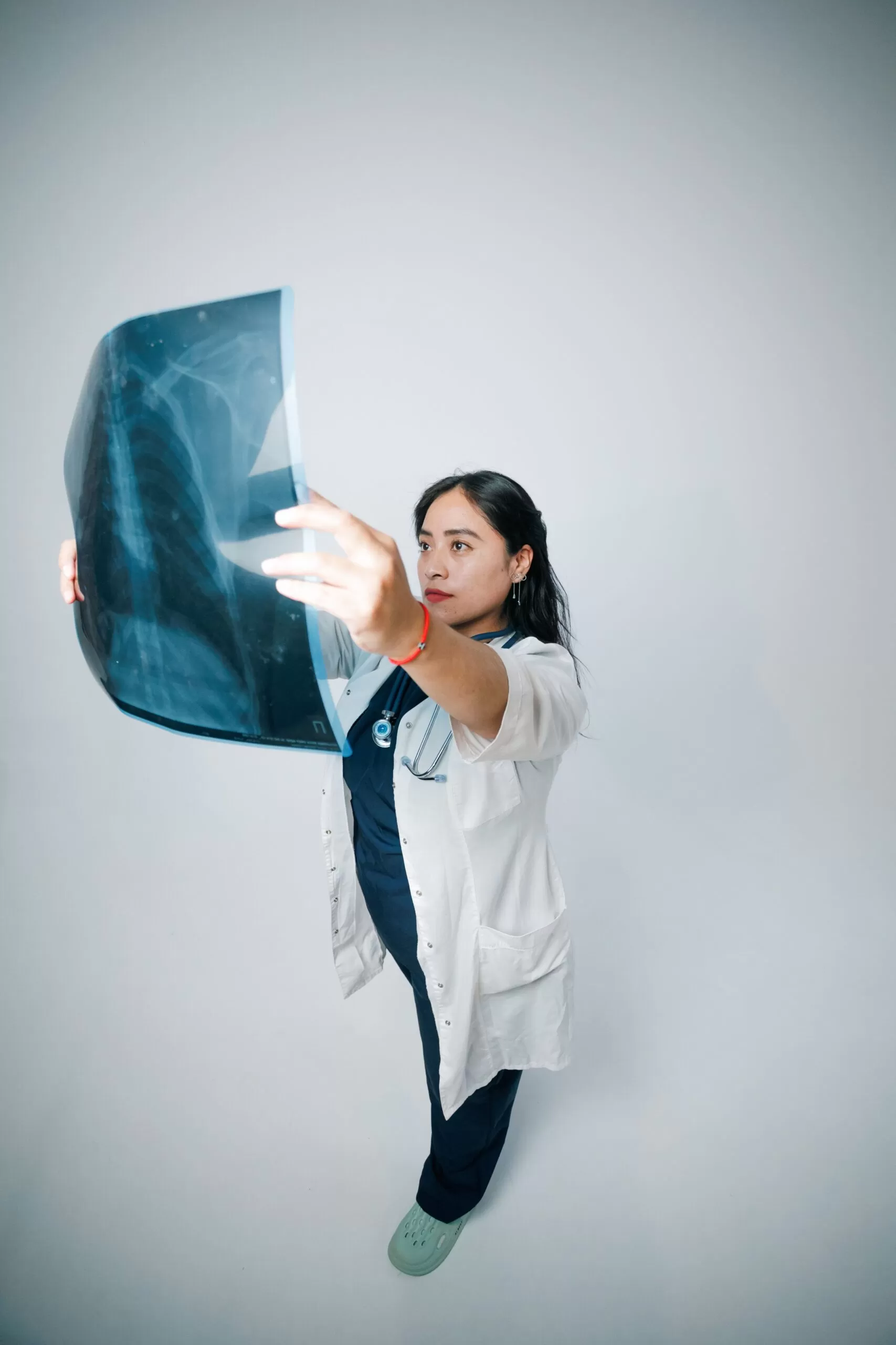 Medical professional in a white coat holding up an X-ray film to examine a chest image against a plain backdrop.