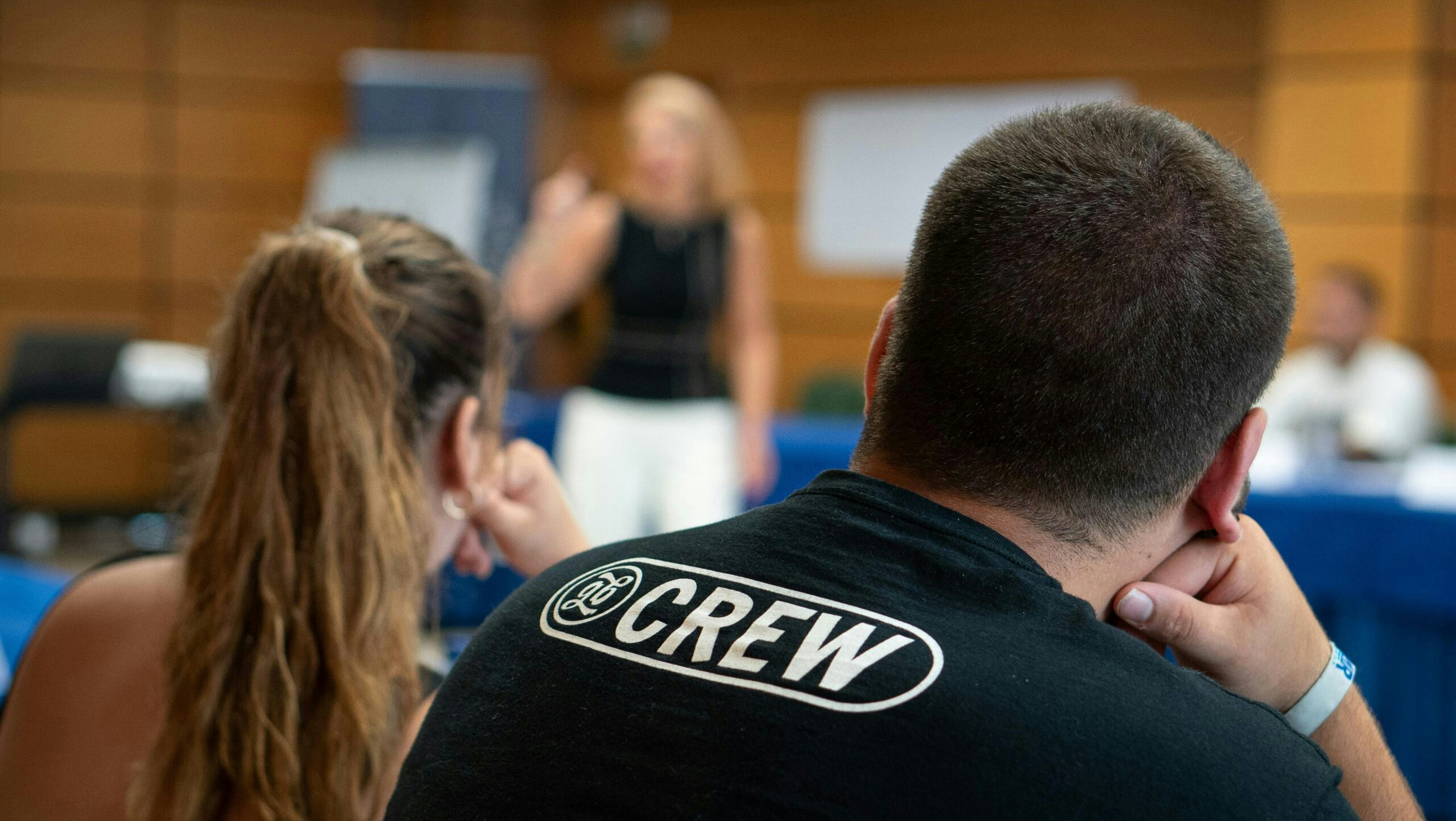 Back view of two people listening in a conference room; the man in the foreground wears a black shirt with a large 'CREW' logo on the back, while a presenter stands blurred at the front.