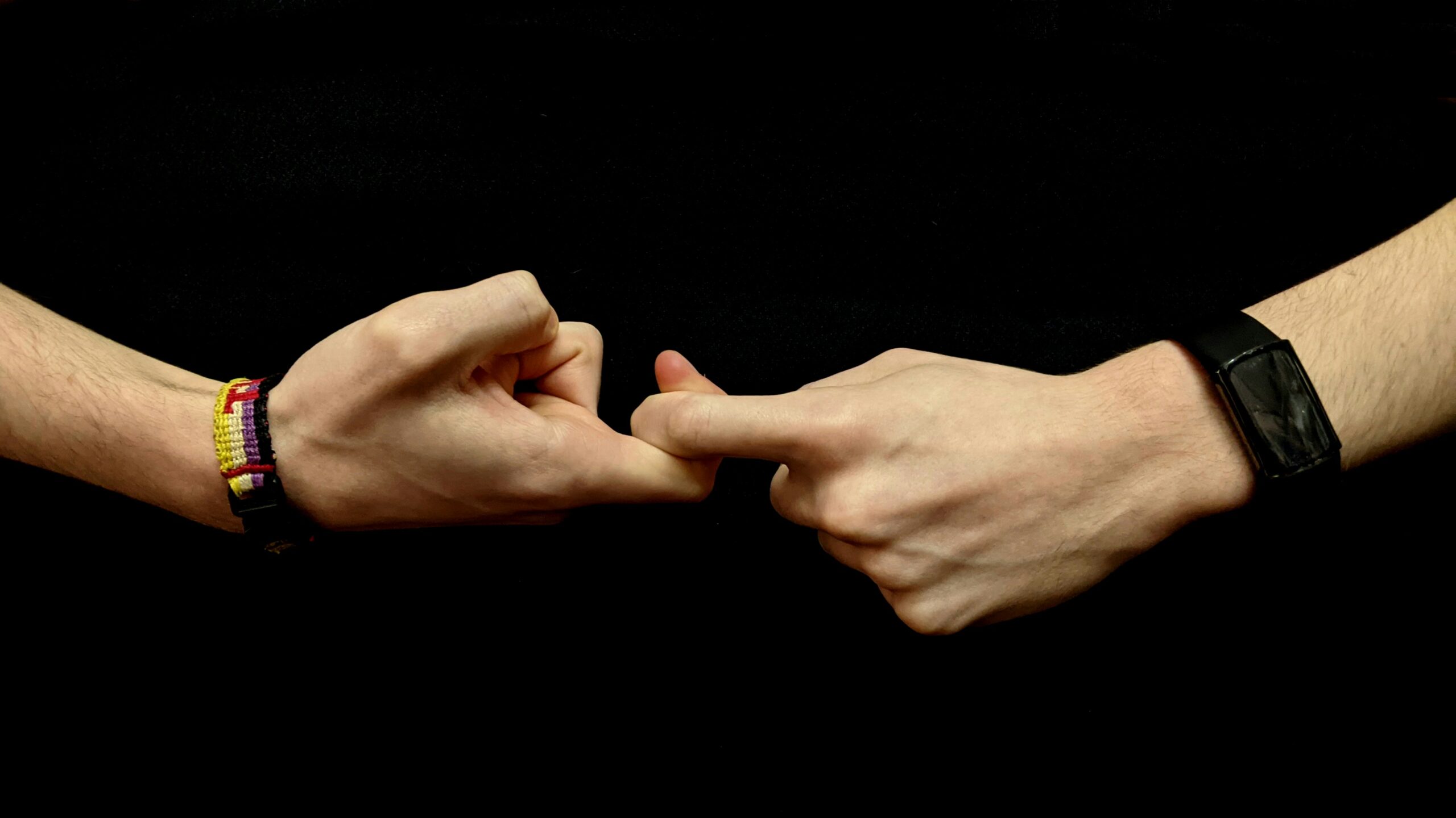 Two people fist-bump against a black background; left wrist has a colorful striped bracelet, right wrist a black smartwatch.