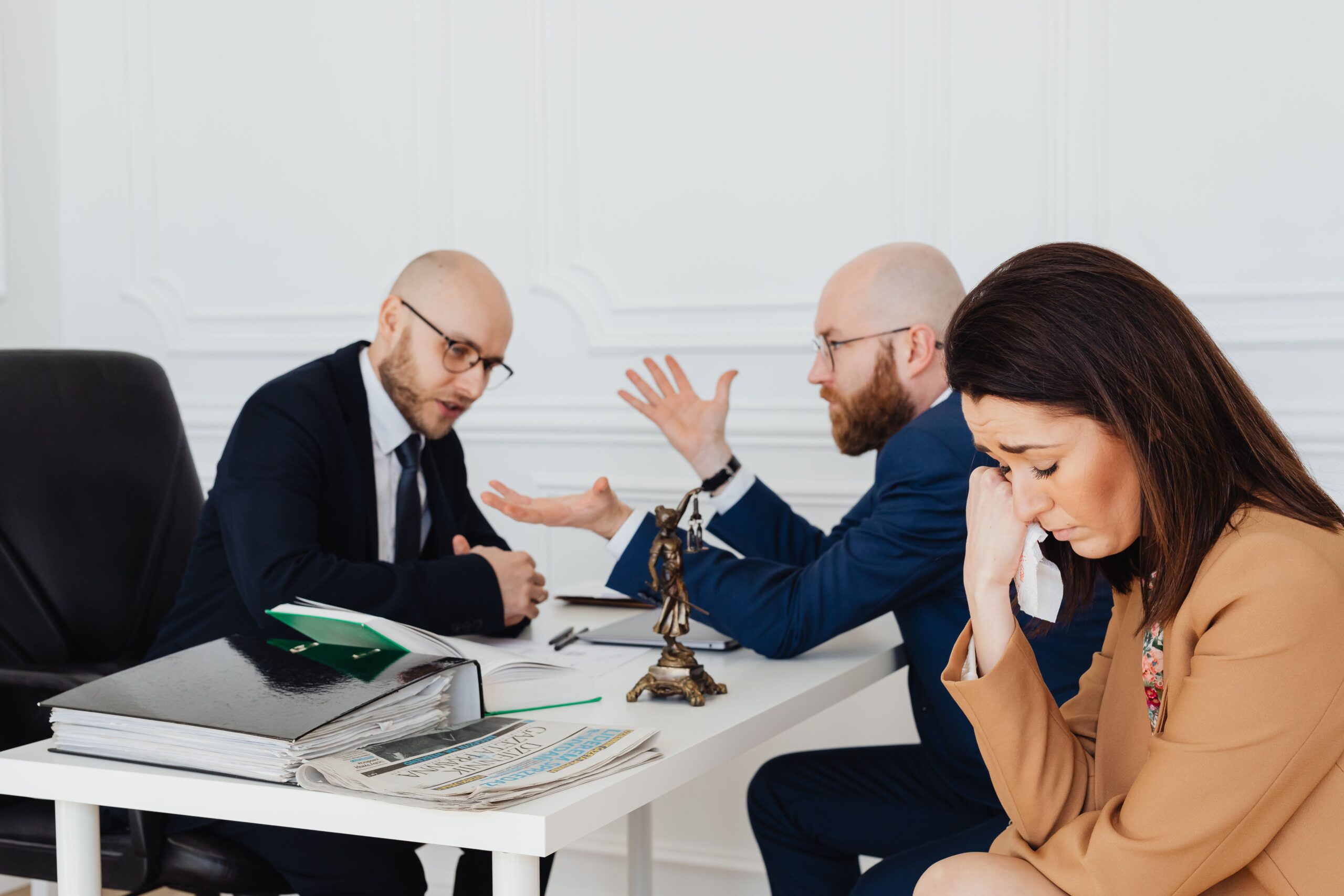 Three coworkers in a conference room: two men in suits argue across the table while a woman on the right looks upset and wipes her eye with a tissue.