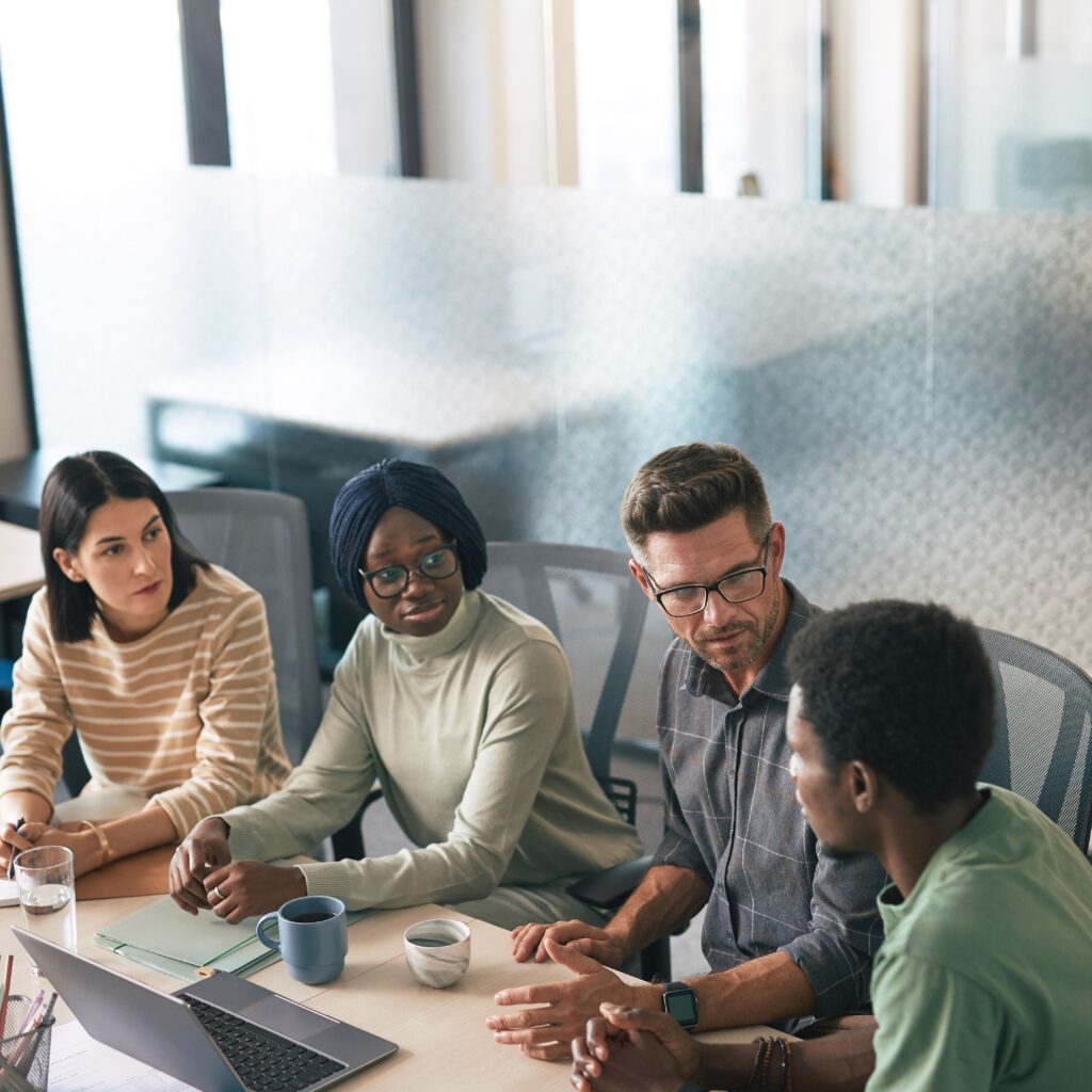 Five colleagues of diverse backgrounds sit around a conference table in a bright office, discussing with notebooks, a laptop, and drinks.