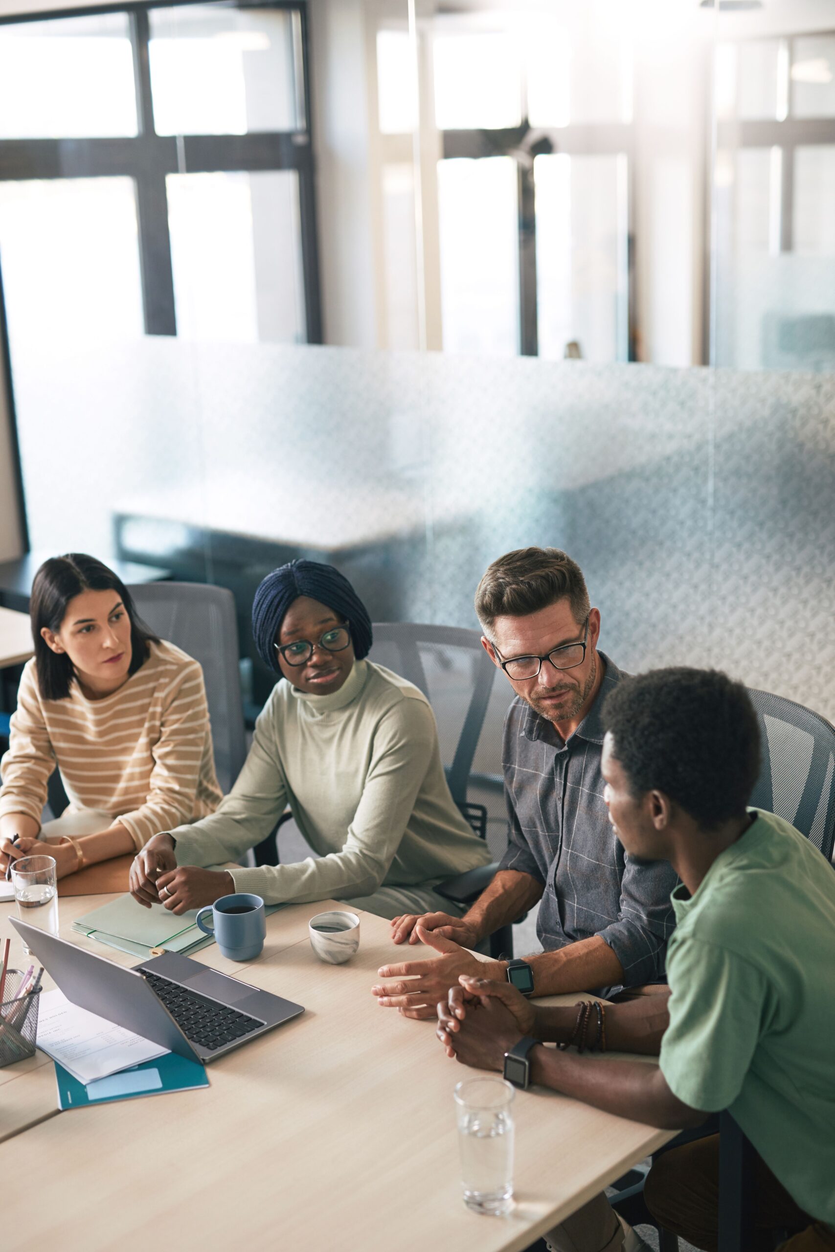 Five colleagues of diverse backgrounds sit around a conference table in a bright office, discussing with notebooks, a laptop, and drinks.