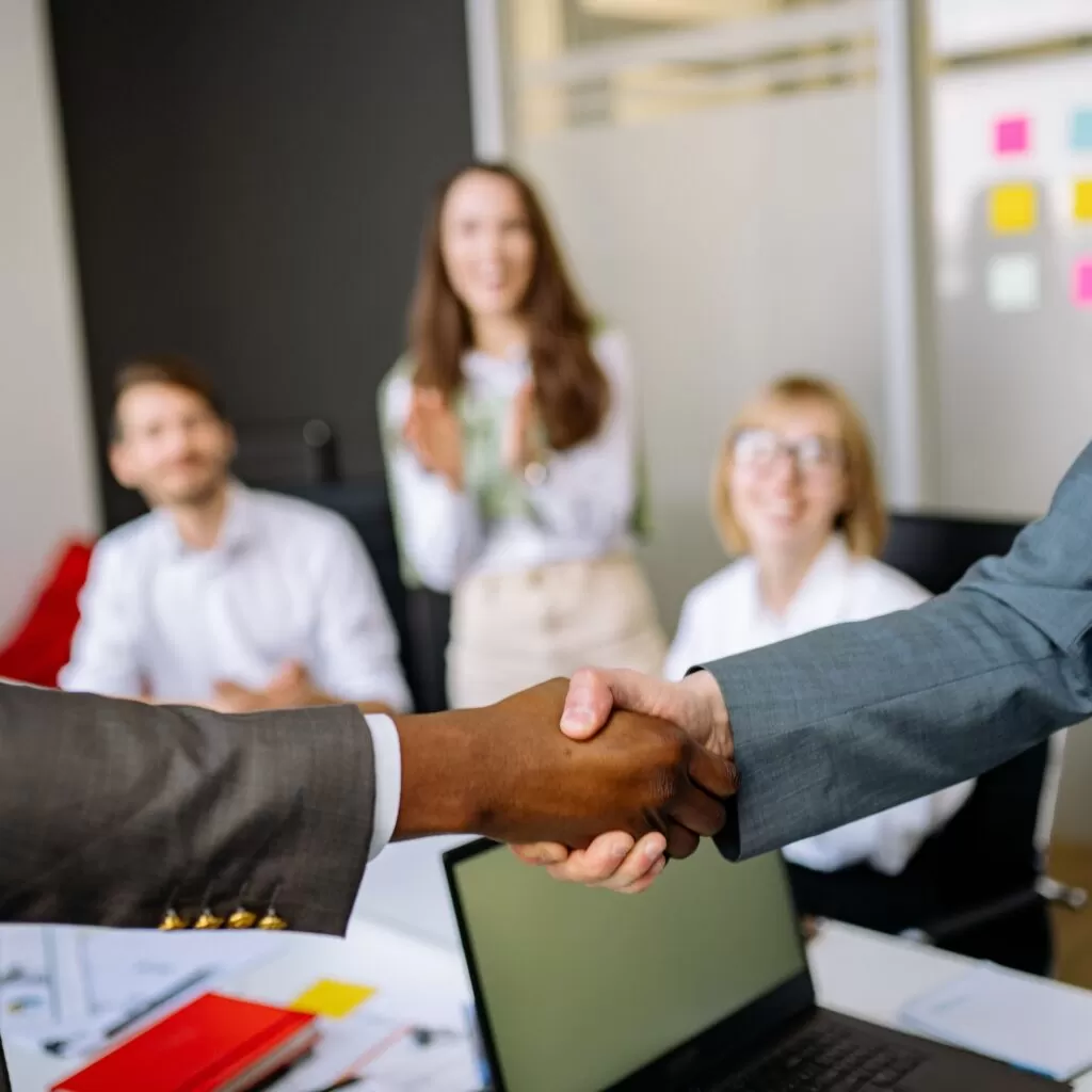 Two professionals shake hands over a desk to seal an agreement, with colleagues blurred in the background in a modern office.