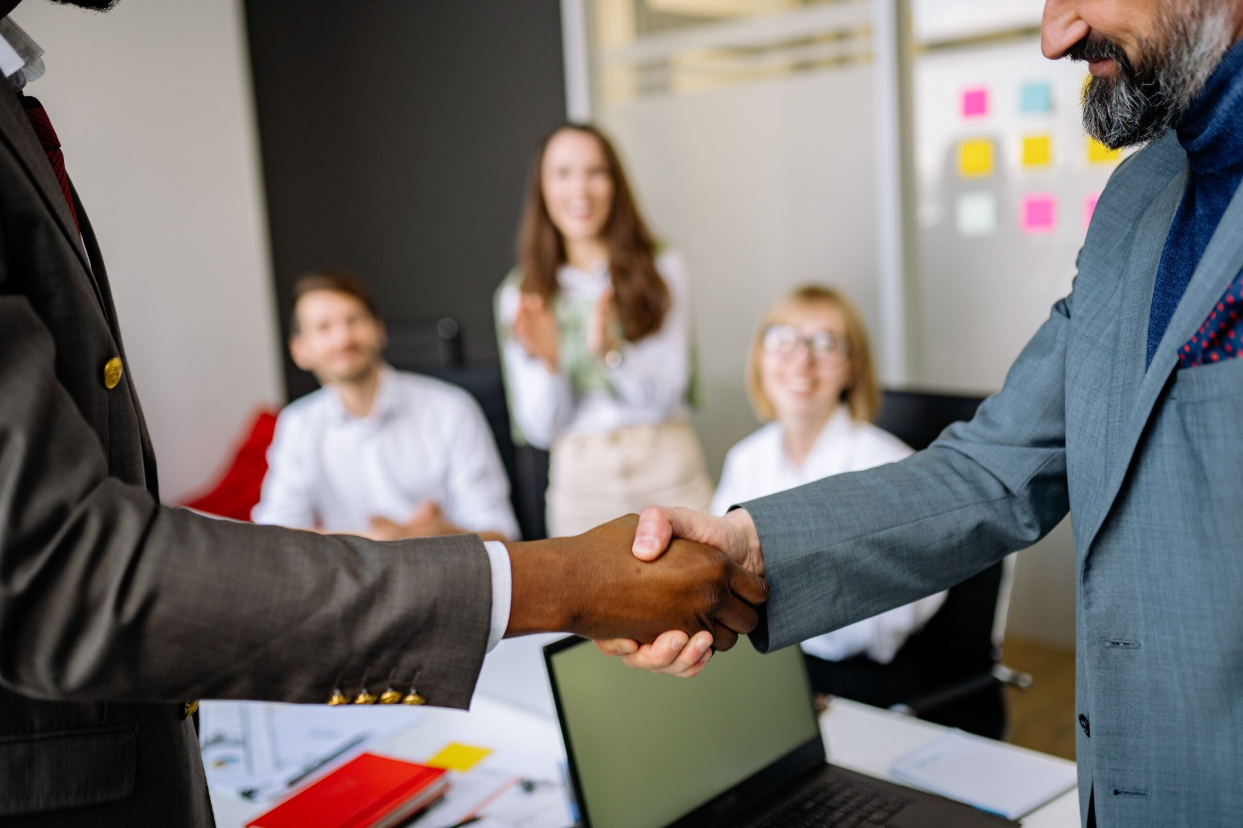 Two professionals shake hands over a desk to seal an agreement, with colleagues blurred in the background in a modern office.