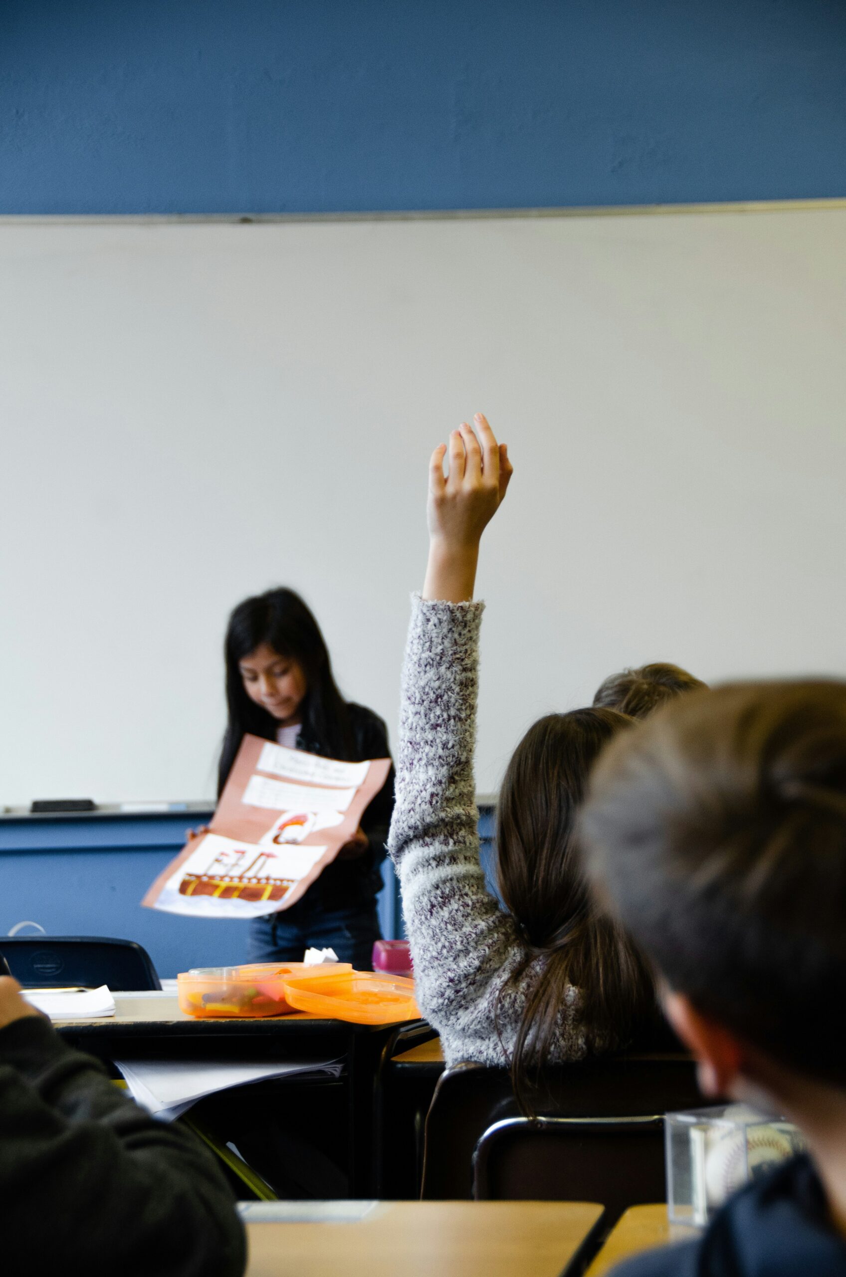 Student raises hand in a classroom as a teacher reads from a booklet in the background.